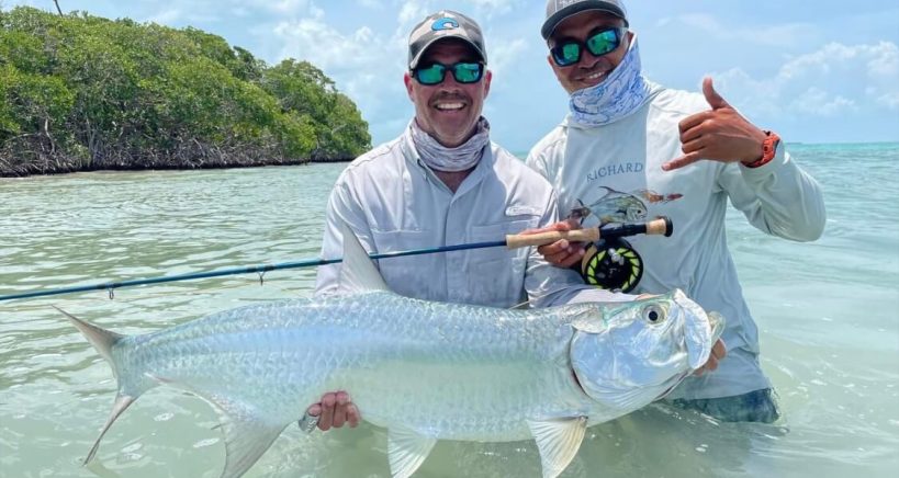 Two anglers standing in caribbean waters holding up a large tarpon fish.