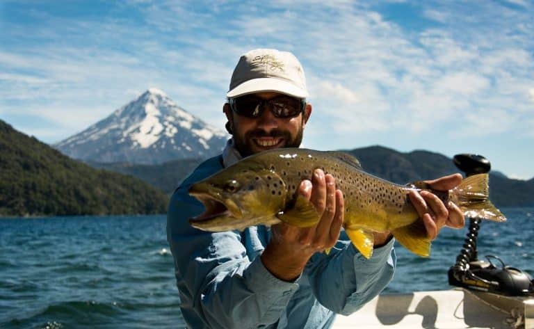An angler holds up an impressive brown trout from a boat out on the open water with a large mountain peak behind them.
