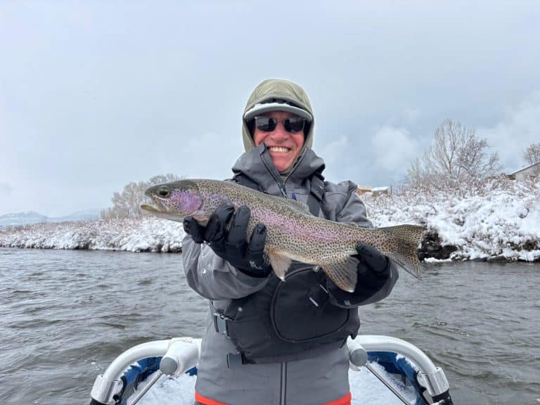 A guest smiles while holding a large rainbow trout on a boat with snow in the background of the river.