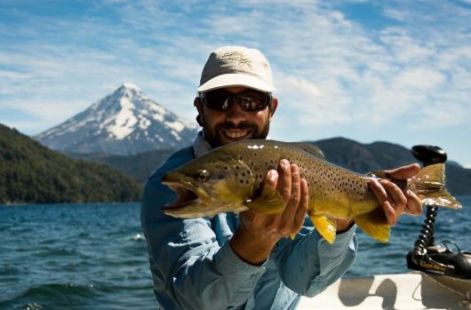 An angler holds up an impressive brown trout from a boat out on the open water with a large mountain peak behind them.