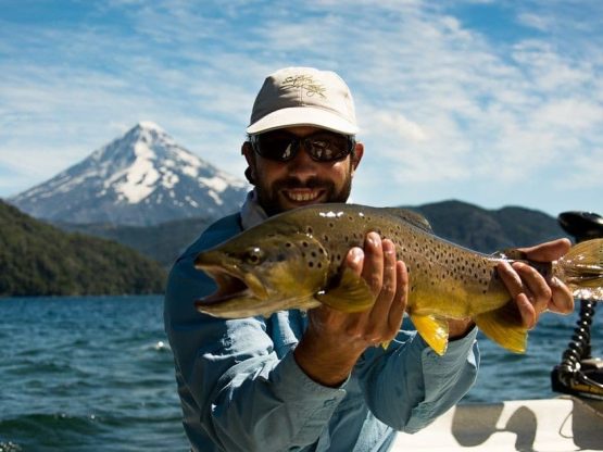 An angler holds up an impressive brown trout from a boat out on the open water with a large mountain peak behind them.