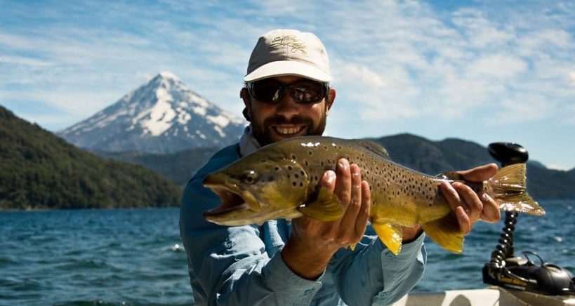 An angler holds up an impressive brown trout from a boat out on the open water with a large mountain peak behind them.