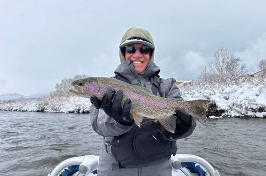 A guest smiles while holding a large rainbow trout on a boat with snow in the background of the river.